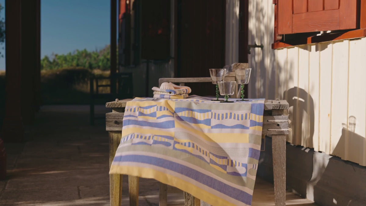 Nappe à rayures jaunes et lavande soulevée par le vent sur une table en bois, devant une maison aux volets rouges